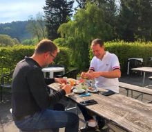 Zwei Männer sitzen draußen an einer Holzgarnitur und vespern 