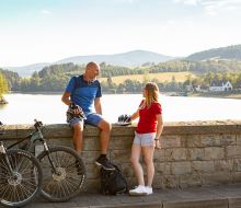 Ein Mann sitzt auf der Staumauer, eine junge Frau steht davor, im Hintergrund der Diemelsee